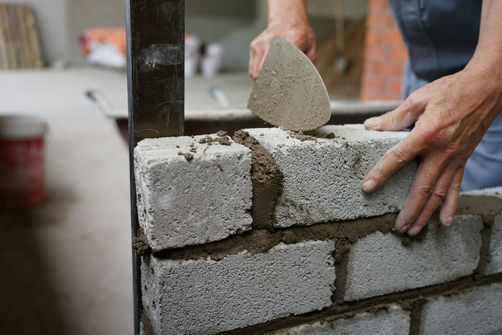 Hands setting a concrete block into fresh mortar, a trowel raised above it