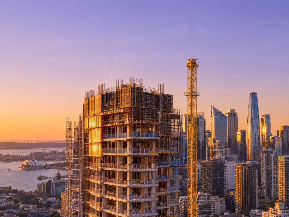 A tower under construction beside a yellow tower crane at sunset, with the Sydney skyline behind