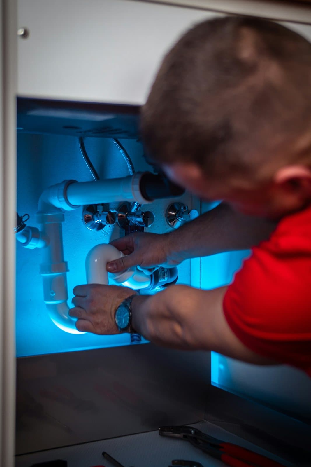 A plumber fitting the waste pipes under a sink, inside a cupboard lit blue