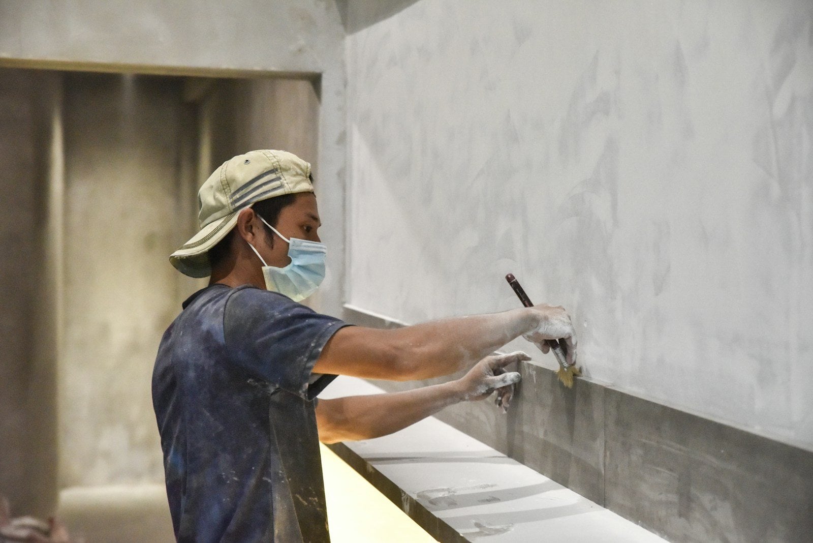 A worker in a face mask brushing a grey coating along the base of a wall